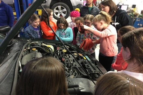 Inspiring Girls to Reach for the Stars in the Automotive World in South Burlington, VT Girlington Garage owner Demeny Pollitt guiding young girl scout troup on how to check oil on car in shop