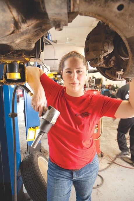 Inspiring Girls to Reach for the Stars in the Automotive World in South Burlington, VT image of Girlington Garage owner Demeny Pollitt standing confidently underneath car chassis while holding auto repair tool 