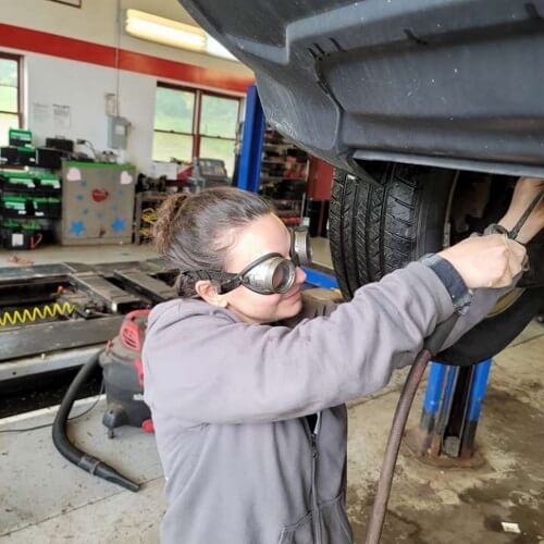 Cha-Ching! Serving Your Car Makes Dollars and Sense | Girlington Garage in South Burlington, VT. Image of a female mechanic doing auto repair and maintenance on a lifted car in Girlington Garage’s auto repair shop.