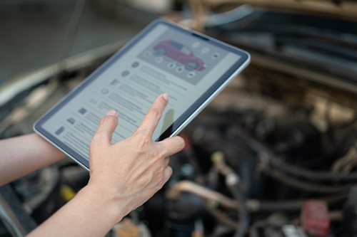 Using the vehicle checklist guideline on the digital tablet during perform preventive maintenance with car engine part as blurred background.